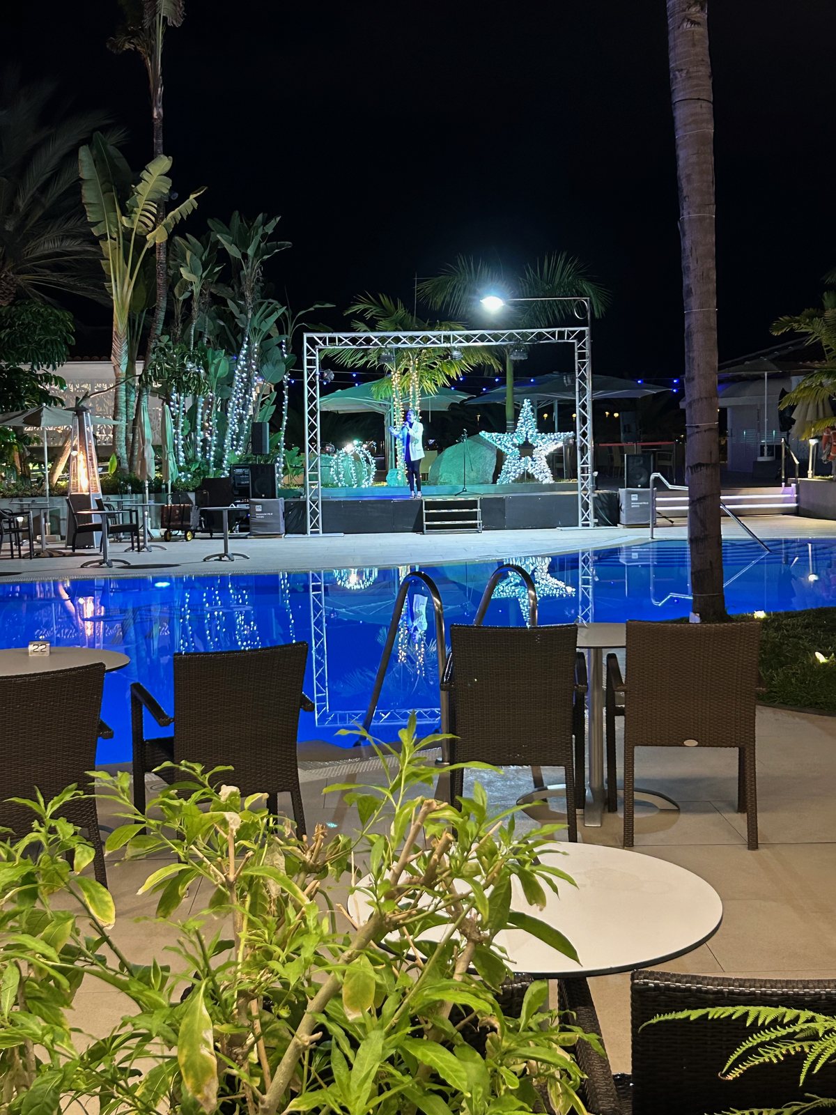 A performer on the outdoor stage across the blue-lit pool, with a patio heater nearby